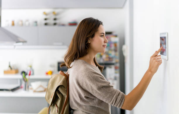 Woman using a smart home control panel on a wall in a modern kitchen to automate home systems.